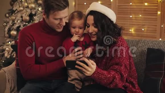 爸爸妈妈和小男孩坐在圣诞树附近的沙发上背景上闪烁着闪闪发光的灯光微笑着看着视频的预览图