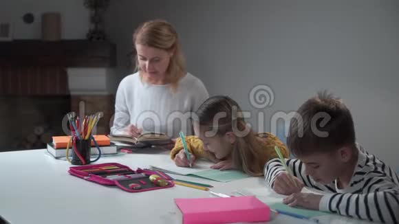 年轻的高加索女人看书检查孩子在练习本中写作男孩和女孩一起做家庭作业视频的预览图