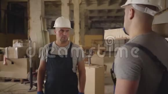 两名戴着白色防护头盔和蓝色制服的白种人建筑工人站在施工现场交谈视频的预览图