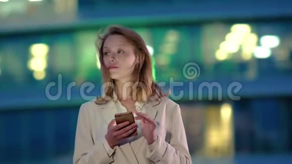 一个金发女人在商务楼附近拿着智能手机等着打电话视频的预览图