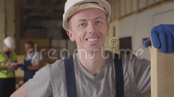 一位戴着白色头盔的白人年轻建筑工人站在木板上微笑的肖像他的同事在谈论视频的预览图