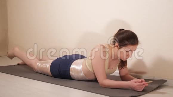 一个女人躺在健身房的垫子上拿着手机大腿和腹部用塑料包裹视频的预览图