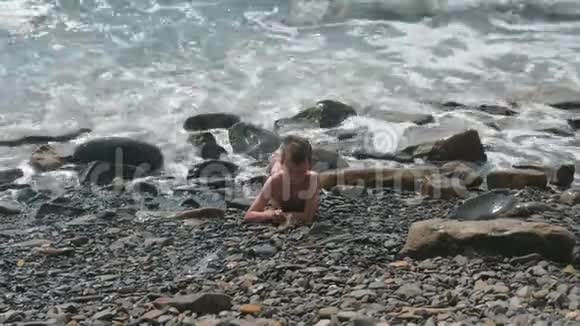 男孩躺在石滩上在海浪中嬉戏视频的预览图