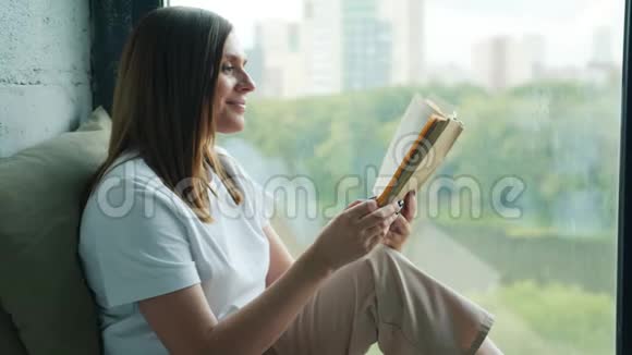 美丽的女人在家中的窗台上缓慢地读着书视频的预览图
