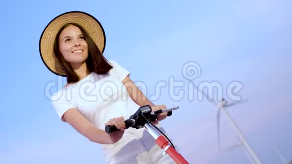 一个年轻的女人戴着草帽旁边有一辆电动滑板车和一台风力发电机视频的预览图