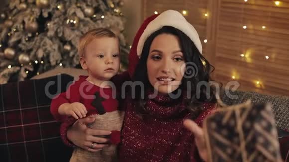 靠近微笑美丽的妈妈戴着桑塔帽抱着她的小男孩坐在客厅附近的沙发上视频的预览图