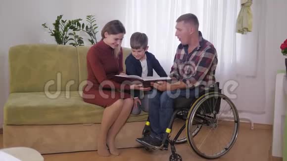 家庭在家幸福的家庭和坐轮椅的父亲一起在房间里读一本有趣的书视频的预览图