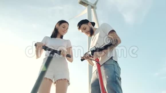 一个男人在附近的风力发电机的背景下向他的女朋友展示了电动滑板车的控制装置视频的预览图