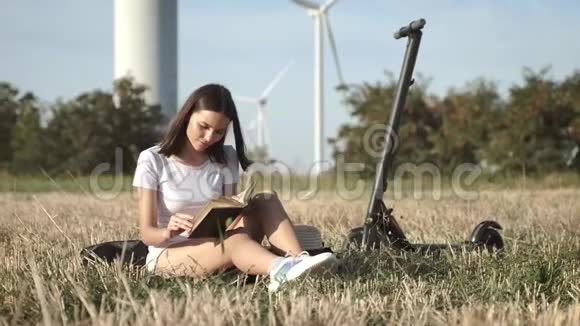一个女孩坐在电动滑板车附近在草原上阅读一本书背景是风力发电站视频的预览图
