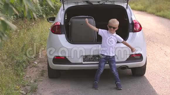 快乐的小男孩玩得很开心在带手提箱的汽车后备箱旁边跳舞视频的预览图