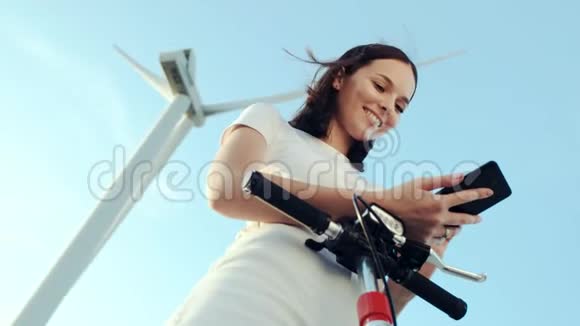 这个女孩在她的电动滑板车旁边使用一部智能手机背景是风力发电机视频的预览图