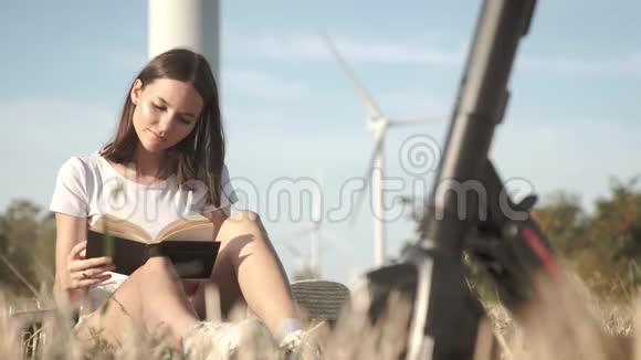这个女孩在风电场的背景下在她的电动滑板车旁边的露天看一本书视频的预览图