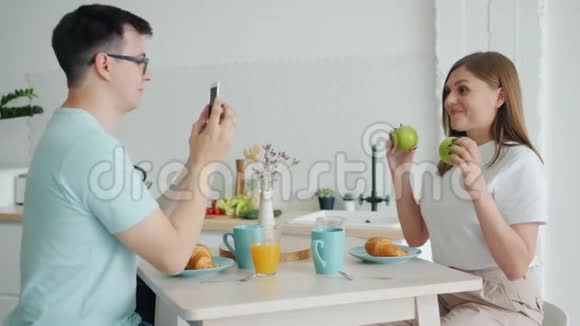 女孩拿着水果摆姿势而男孩拿着智能手机在餐桌上拍照视频的预览图