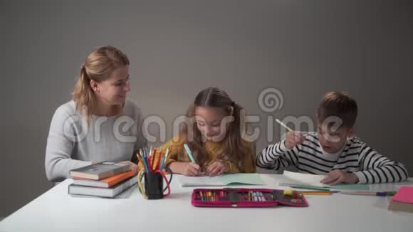 聪明的高加索母亲和两个孩子坐在桌子旁做家庭作业的肖像姐妹和兄弟视频的预览图