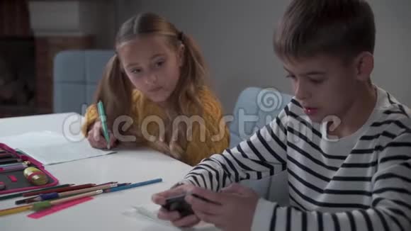 白人男孩在学校或家里玩智能手机游戏他的同学或姐姐在做作业或任务视频的预览图