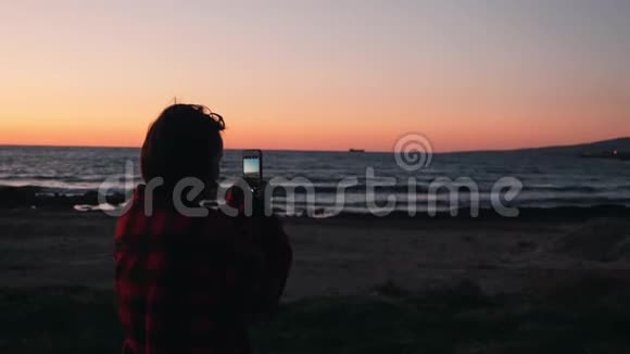 后观年轻女性站在海滩上制作海上日落的照片和视频女人为美丽的日落拍照视频的预览图