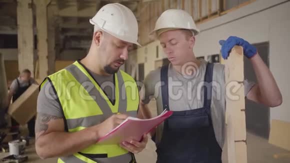 两名成年男性白种人建筑工人站在建筑工地上交谈穿着绿色背心的胡子男视频的预览图