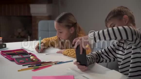 白人男孩厌倦了在学校玩智能手机他的同学或姐姐坐在他旁边学习视频的预览图