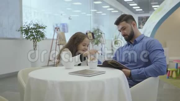 富有的白种人男人和小女孩坐在机场贵宾等候区看着照片喝着茶或视频的预览图