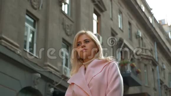 一个年轻漂亮的金发女郎穿着粉红色的外套站在古老建筑的背景下视频的预览图