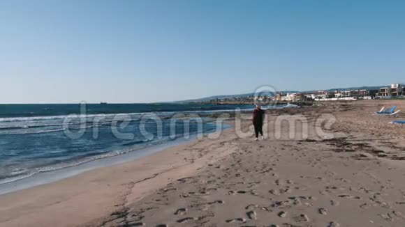 穿着格子红色衬衫的年轻时髦女人沿着空荡荡的海滩散步在多风的秋天天气里孤独悲伤的女人走在岩石海岸上视频的预览图
