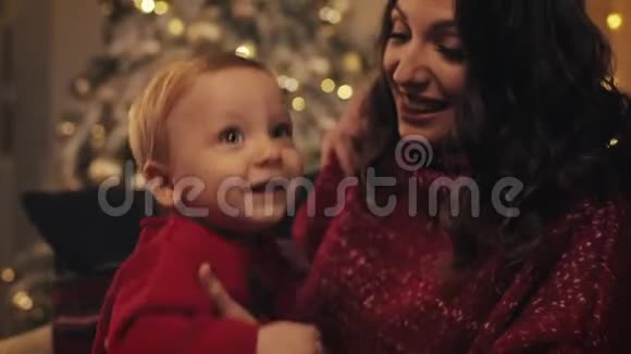 在家庭节日的背景下把可爱的微笑的小男孩抱在妈妈的膝盖上跳圣诞树视频的预览图