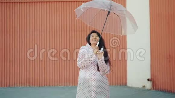 穿着雨衣在户外跳舞拿着雨伞看镜头的快乐亚洲女孩视频的预览图