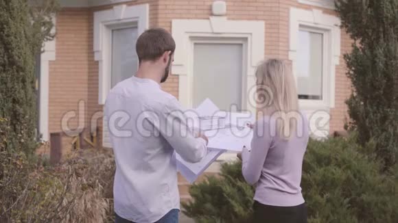 成年英俊的白种人建筑师在新建的房子前展示年轻女子的蓝图客户和视频的预览图