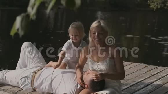 年轻幸福的家庭爸爸妈妈和儿子在河边的码头上阳光明媚的一天男人躺在女人腿上视频的预览图