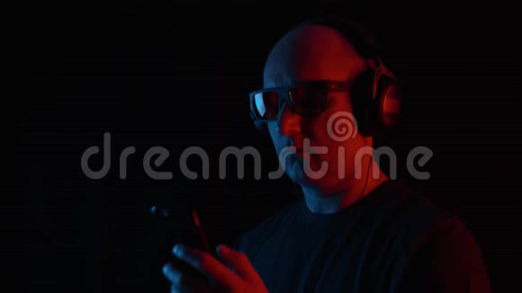 成年男子在黑色背景下听移动音乐红色和蓝色照明肖像秃头男子使用视频的预览图