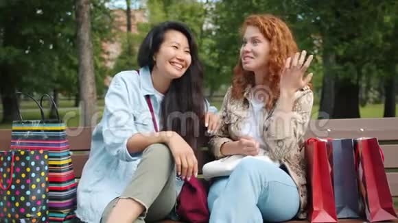美丽的女孩亚裔和高加索人拿着购物袋在公园的长凳上笑视频的预览图