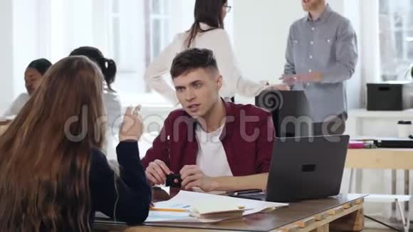 严肃的年轻商人听着女同事的谈话坐在办公室的桌子上在阁楼上时髦地合作视频的预览图