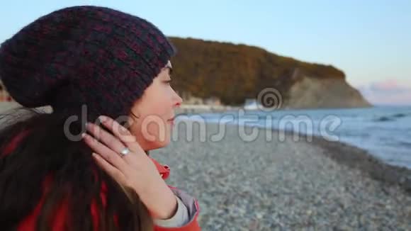 一位留着黑色长发戴着帽子和夹克的漂亮年轻女子在欣赏海景面部特写转过头视频的预览图