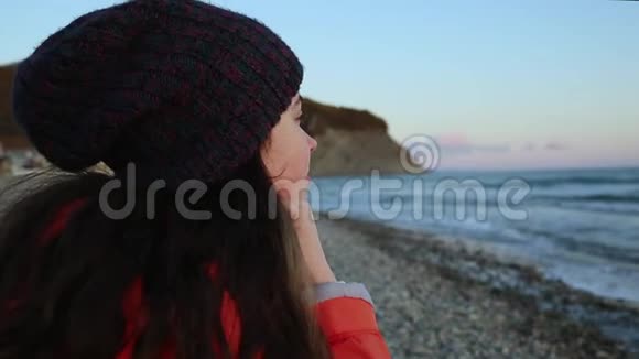 一个漂亮的年轻女人留着长长的黑发戴着帽子和夹克坐着欣赏海浪面部特写视频的预览图