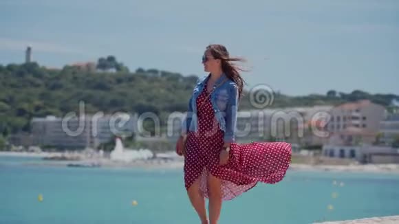 戴着太阳镜穿着浪漫的红色连衣裙和牛仔夹克的快乐的年轻女子走在堤岸的石墙上视频的预览图