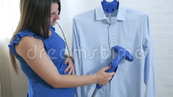 一个年轻的孕妇摸了摸她的胃用蒸汽抚摸男人衬衫蒸衣服的过程视频的预览图