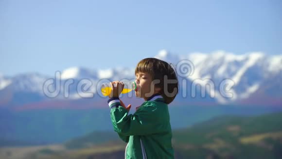 一个非常可爱的男孩在美丽的山脉背景上喝橙汁小男孩在山顶上放松视频的预览图