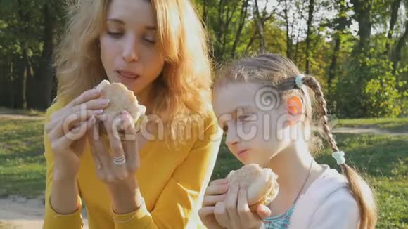 户外照片年轻的金发母亲和她的小女儿坐在公园的长凳上吃芝士汉堡视频的预览图