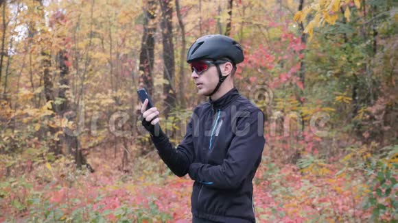 年轻的男运动员穿着自行车服装戴着头盔和眼镜在秋季锻炼前拨打电话和智能手机视频的预览图