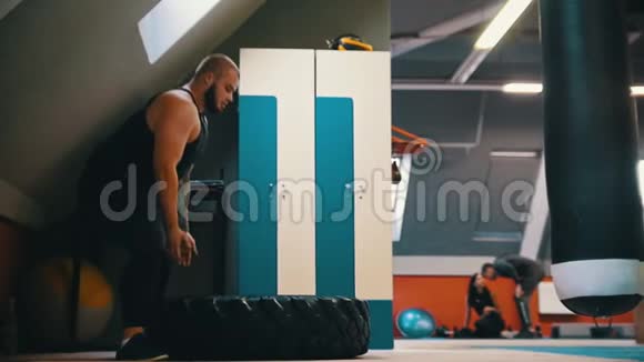 一个长胡子的健美男子在健身房里一遍又一遍地翻着地板上的轮胎另一个人坐在地板上视频的预览图
