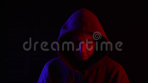 穿着深色帽衫的人在蓝色和红色灯光下的黑色背景上仰望镜头一幅神秘男子的画像视频的预览图