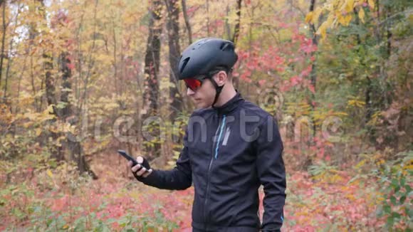 在秋季城市公园的智能手机上自拍的骑自行车服装的年轻迷人的Sports家伙拍摄照片的专业自行车手视频的预览图
