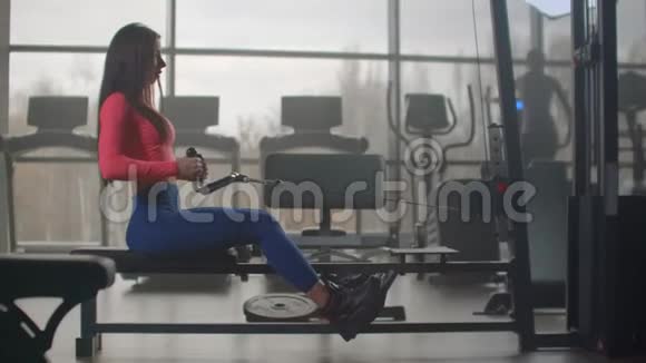 健身房的强化力量训练一位坐着的女士在大型视窗的背景下拉动模拟器的重量视频的预览图