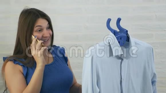 一个年轻的女人在电话里说话用蒸汽抚摸男人衬衫蒸衣服蒸的过程蓝色视频的预览图