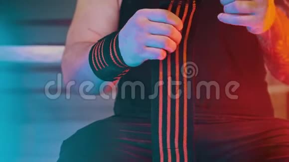 一个肌肉发达的男人双手上有肌肉手腕上有弹性绷带坐在健身房里戴着霓虹灯视频的预览图