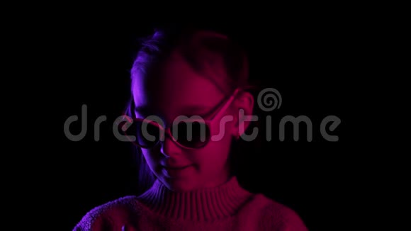 戴着太阳镜的微笑少年女孩在黑暗的工作室里用霓虹灯照相机戴眼镜的年轻女孩视频的预览图