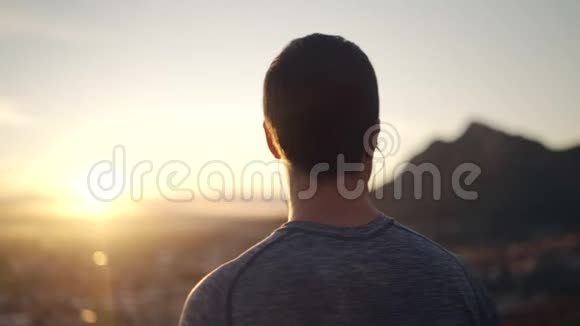 一位戴着耳机的男性运动员的侧景从山区快乐的人那里欣赏早晨的阳光和城市景观视频的预览图
