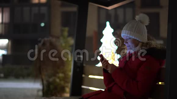 一位美丽的金发美女在一条装饰着冬天街道的长凳上穿着智能手机购物视频的预览图