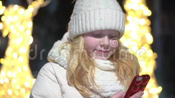 一个美丽的金发女人在冬天装饰着圣诞节的街道上的智能手机里聊天视频的预览图
