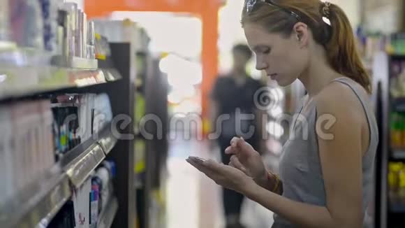 年轻的白种人女人正在美容店里选择持有智能手机的化妆品视频的预览图